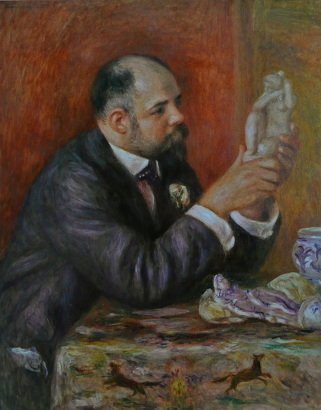 A bearded, balding man in a dark suit seated at a table, examining a small statuette in his hands, in warm red, violet, and ochre tones