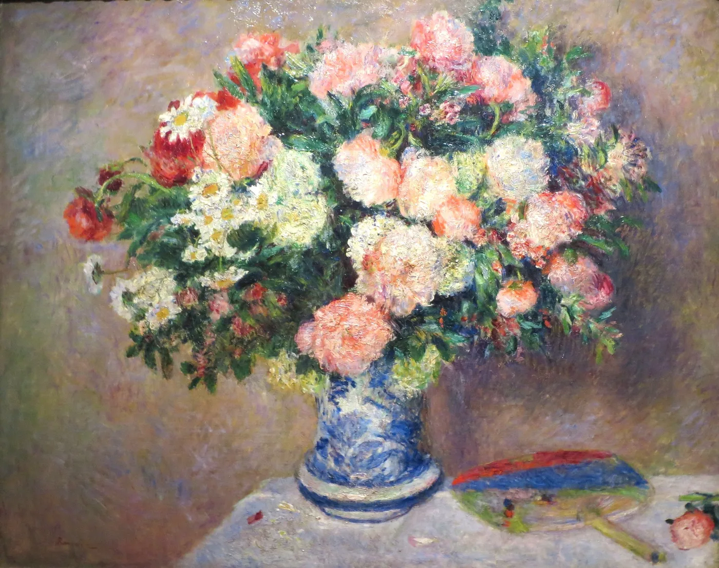 Pink, white and red chrysanthemums overflow a blue and white vase beside a folded Japanese fan on a white cloth