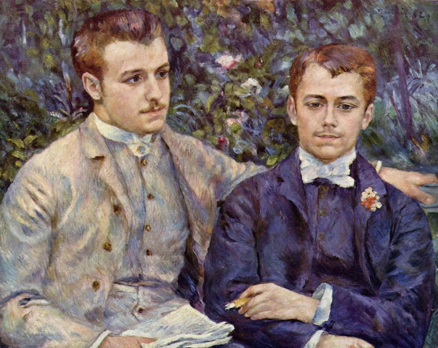 Two young men in suits seated in a garden, one in beige and one in navy blue, against dense green foliage