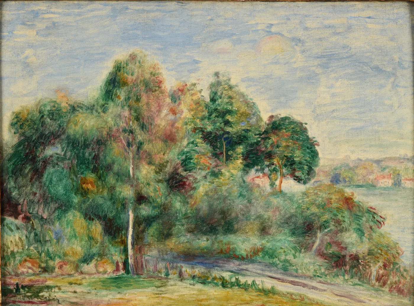 Trees line a road above a river bay, a distant village visible across the water under a pale blue sky.