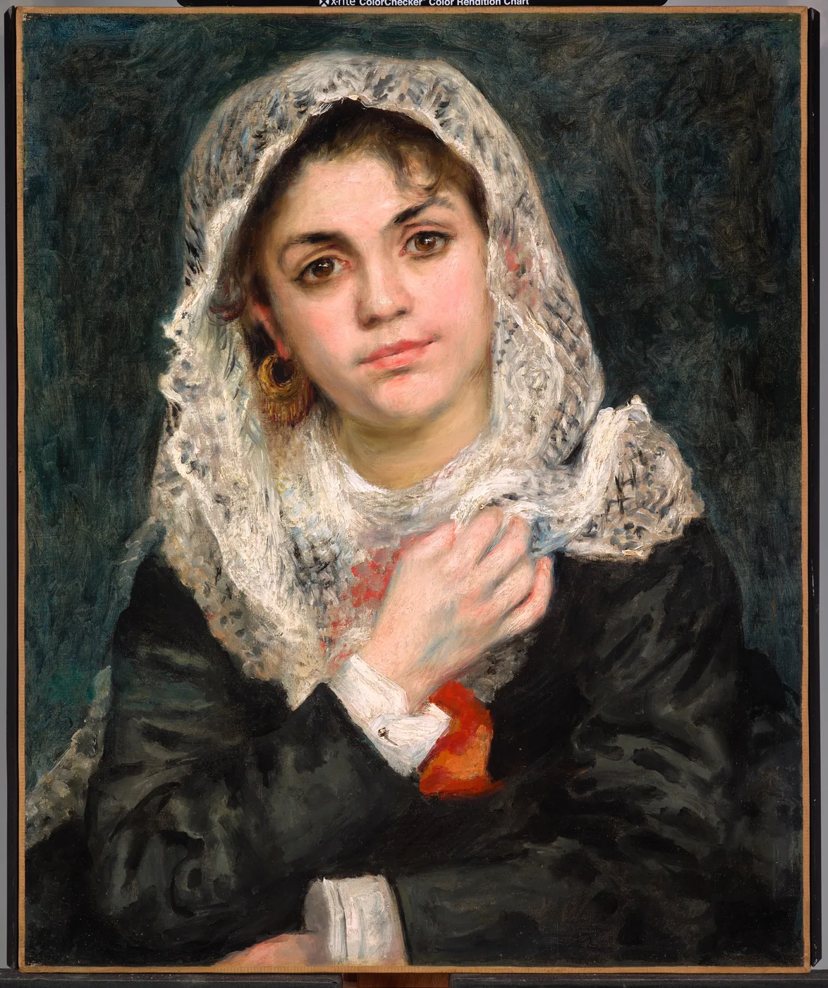 A woman in a white lace shawl draped over her head holds it closed at her neck, gold hoop earrings, against dark green.