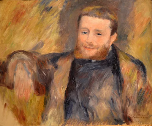 A smiling red-bearded man leans his arm on a chair, wearing a dark scarf against a warm gold background.