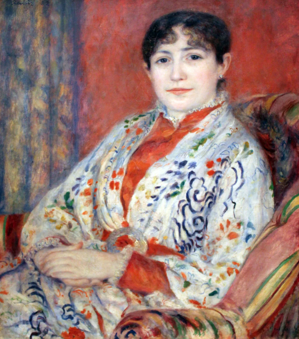 A seated woman in a white kimono-style robe patterned with blue and orange holds a fan, against a red background.