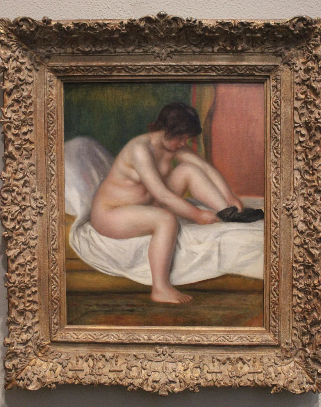 A nude woman with dark hair sits on a white sheet pulling on a black stocking, against green and pink.