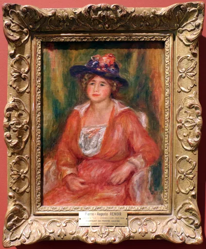 A seated woman in a dark flowered hat and coral-red dress with a lace bodice rests her hands in her lap, against a green backdrop, in coral and navy tones.