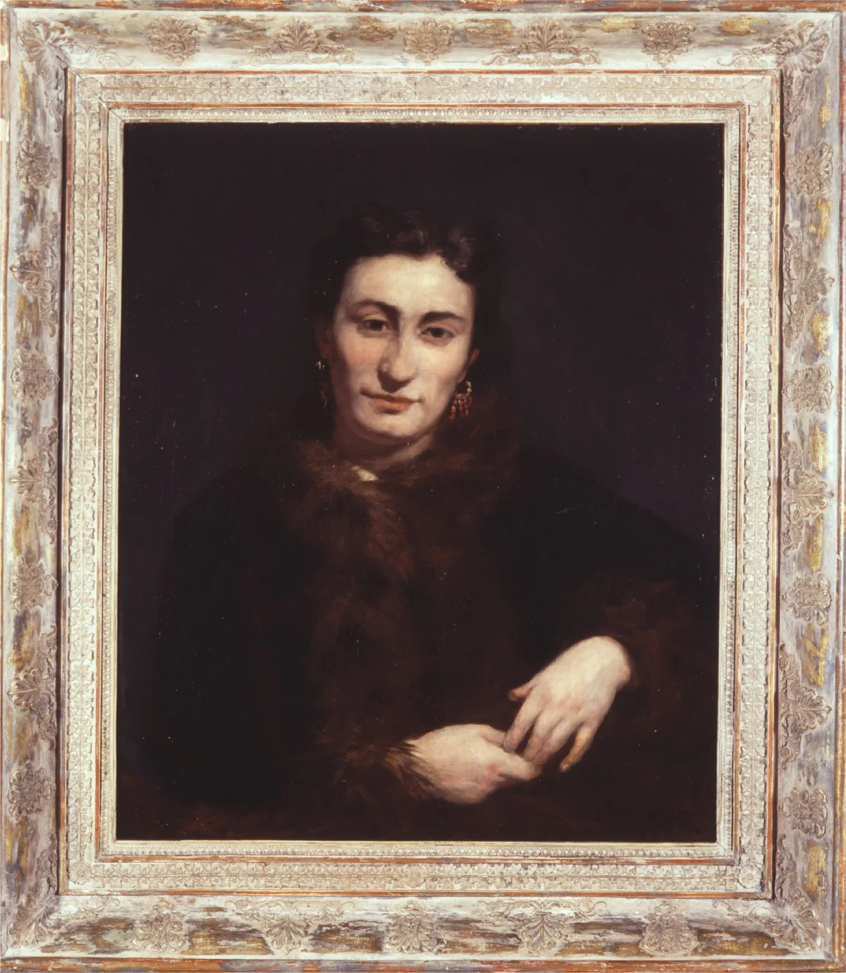 A dark, shadowy portrait of a woman with dark hair and drop earrings, wearing a fur-trimmed coat with one hand resting at her waist.