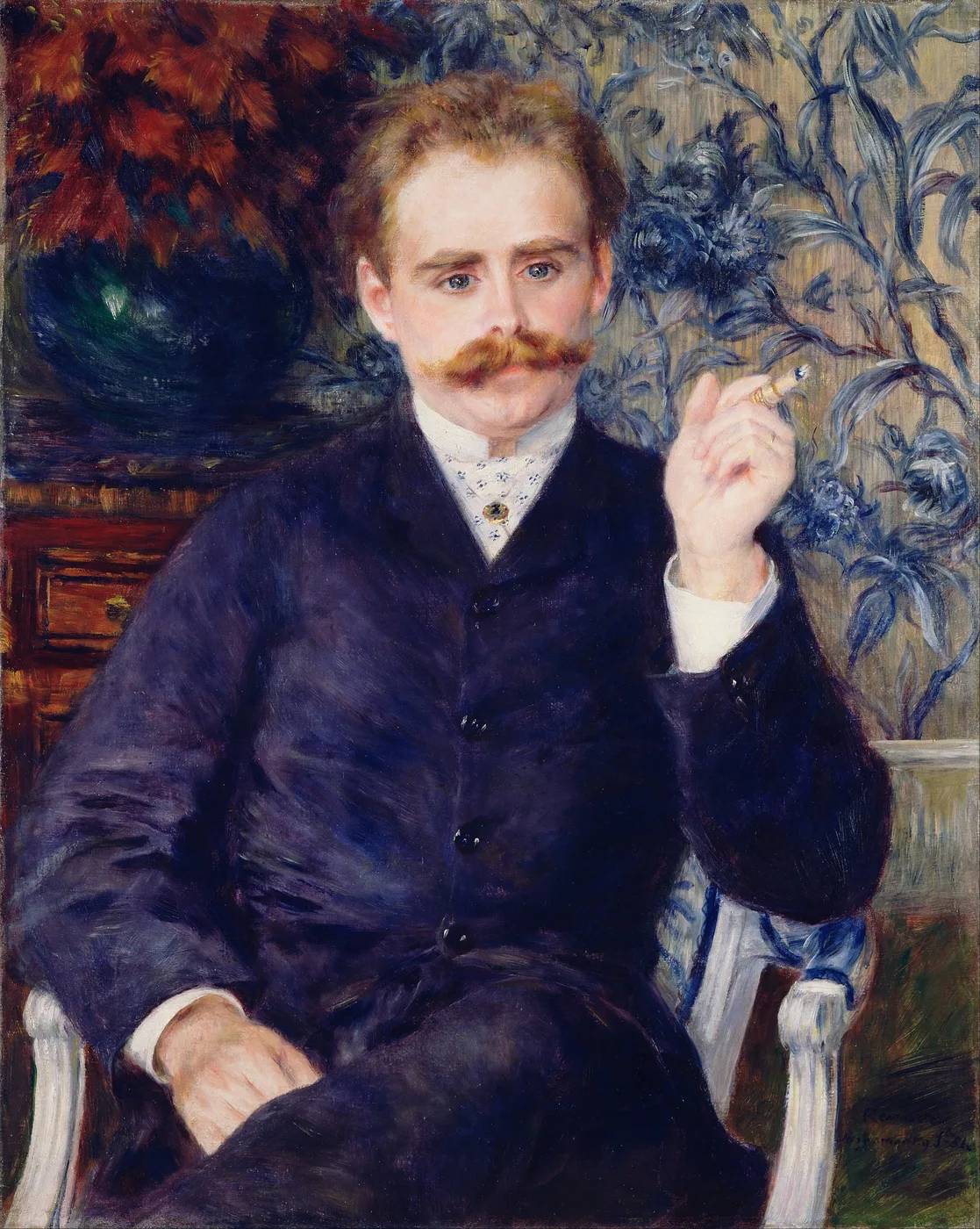 A moustached man in a dark blue jacket seated in a white armchair, holding a cigarette, before floral wallpaper and a vase of red flowers