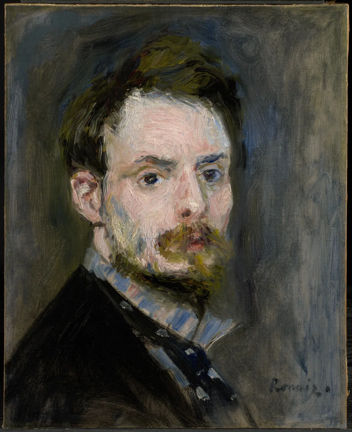 A young bearded man with tousled brown hair looks intently at the viewer, wearing a dark coat over a striped blue shirt, against a grey background
