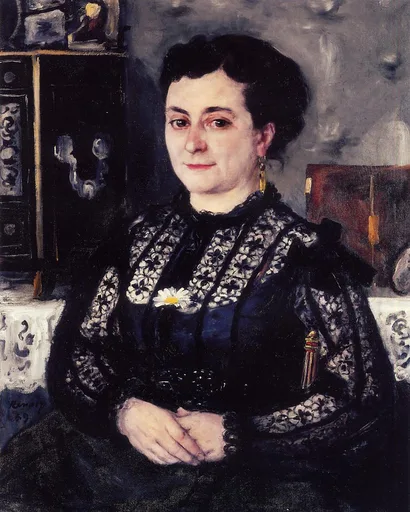 A seated woman with dark hair in a black lace blouse with a white daisy, hands clasped, against a grey wall
