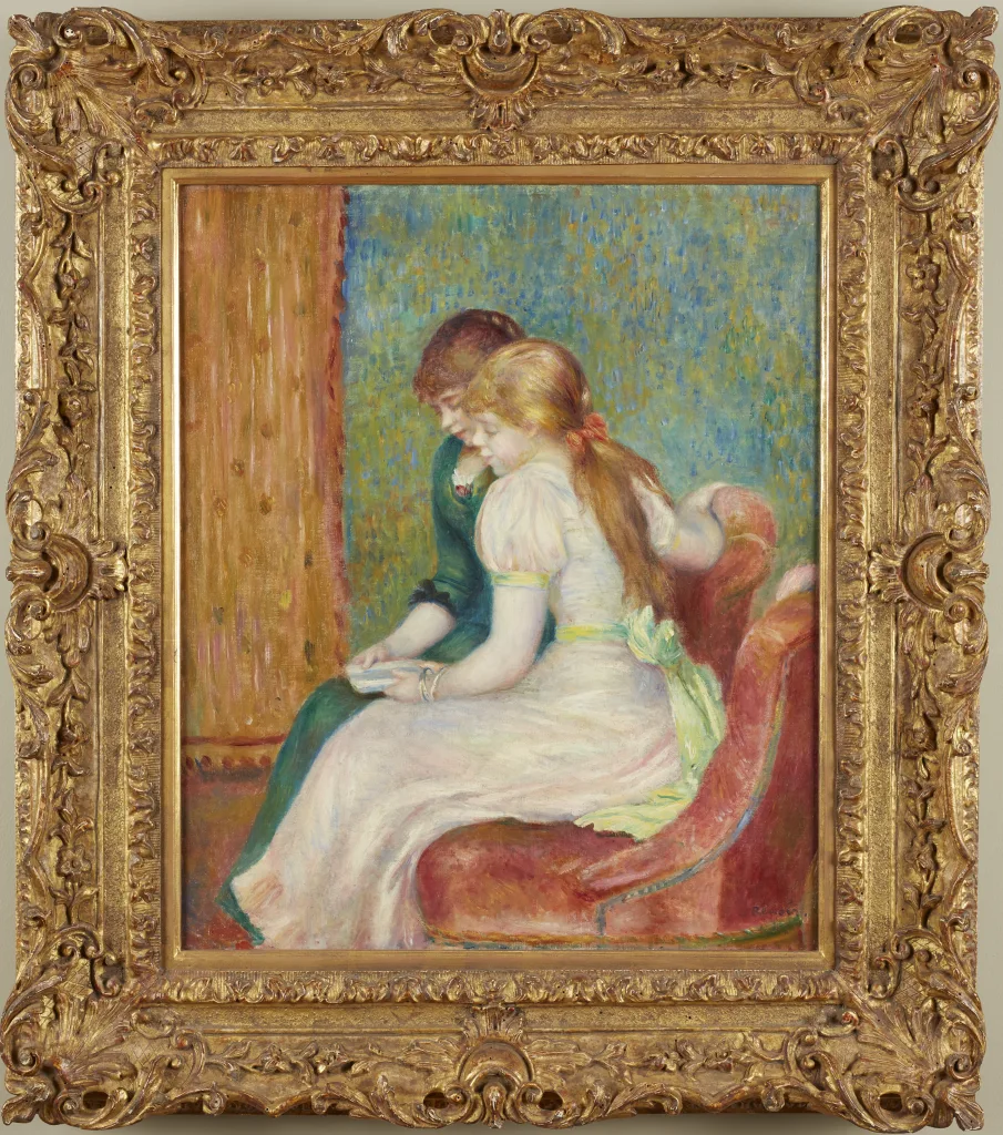 Two young girls seated close together reading on a pink upholstered sofa, in white, green, and soft blue